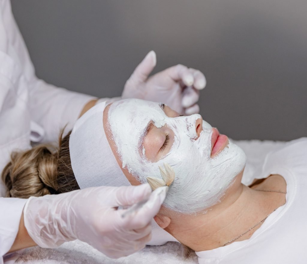 A person is receiving a facial treatment with a white mask applied, while another person is using a brush to apply it.