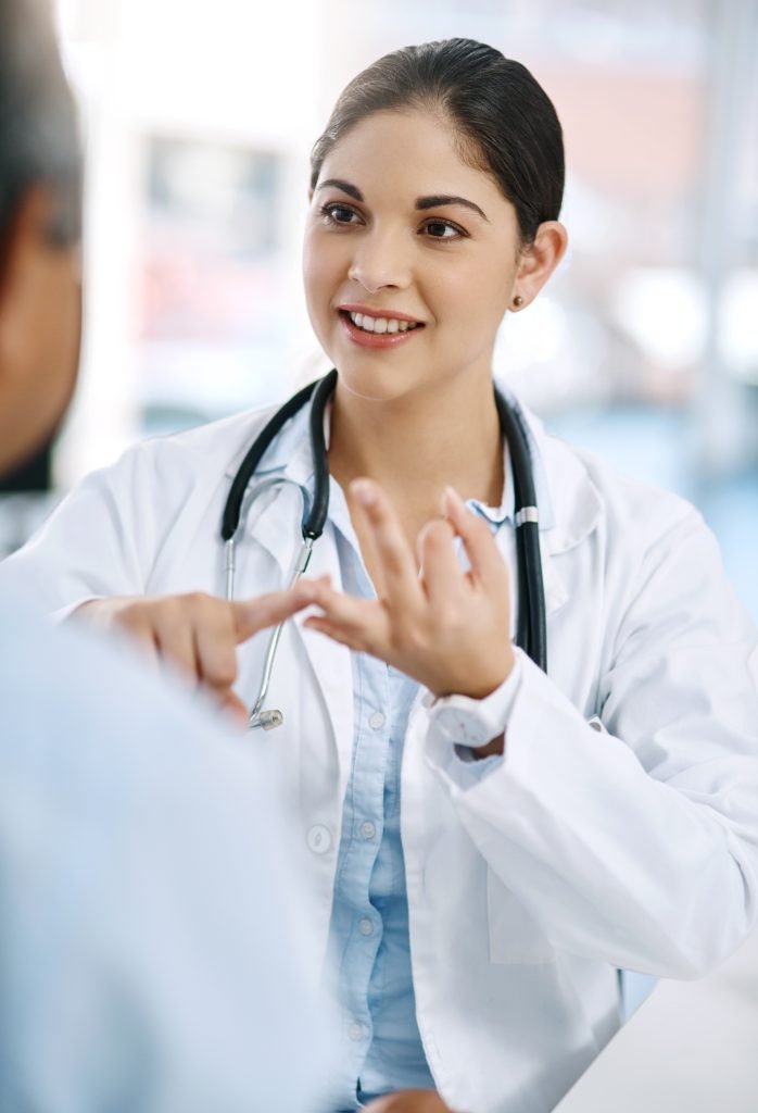 A person in a white coat, wearing a stethoscope, gestures while speaking with another person indoors in a bright, clinical setting.