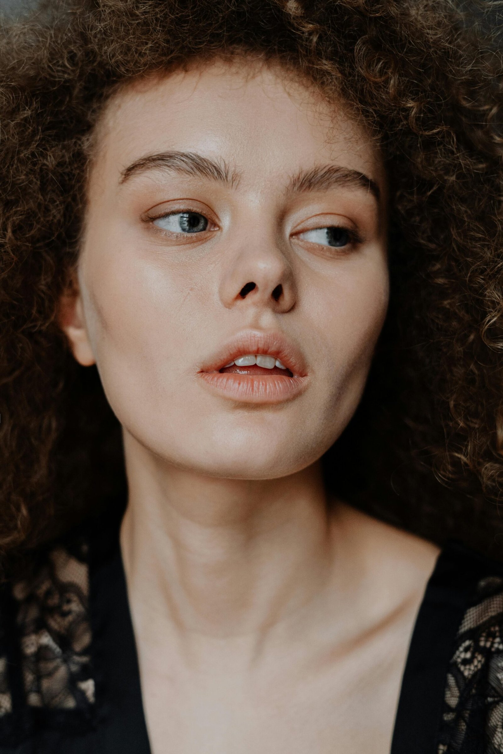 Close-up of a person with curly hair, looking to the side. Wearing black attire with lace detail. Neutral background.