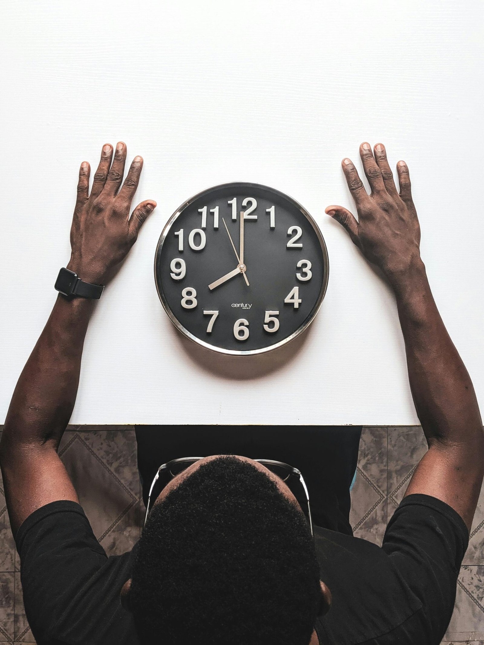 A person sits at a table with a round black clock, hands placed parallel on either side, against a white background.