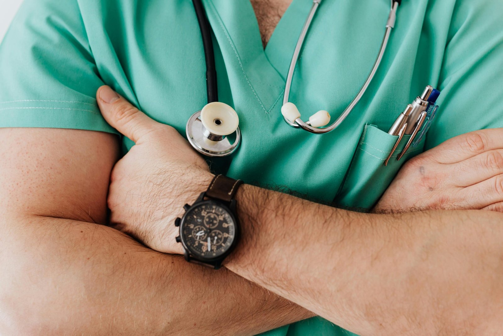 Person in green scrubs with stethoscope and pens, arms crossed, wearing wristwatch. Focus on medical attire, suggesting healthcare professional.