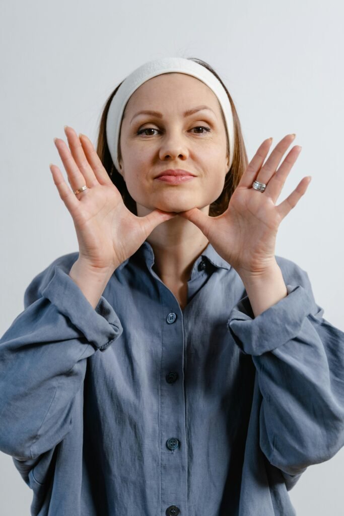 A person wearing a blue shirt and headband poses with hands near face against a neutral background, showcasing rings on fingers.