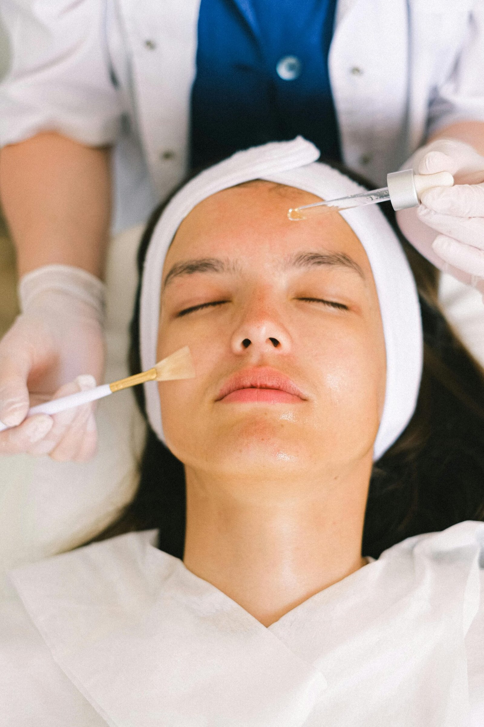 A person receives facial treatment with a brush and dropper while wearing a headband. Professional wearing gloves assists in a spa setting.