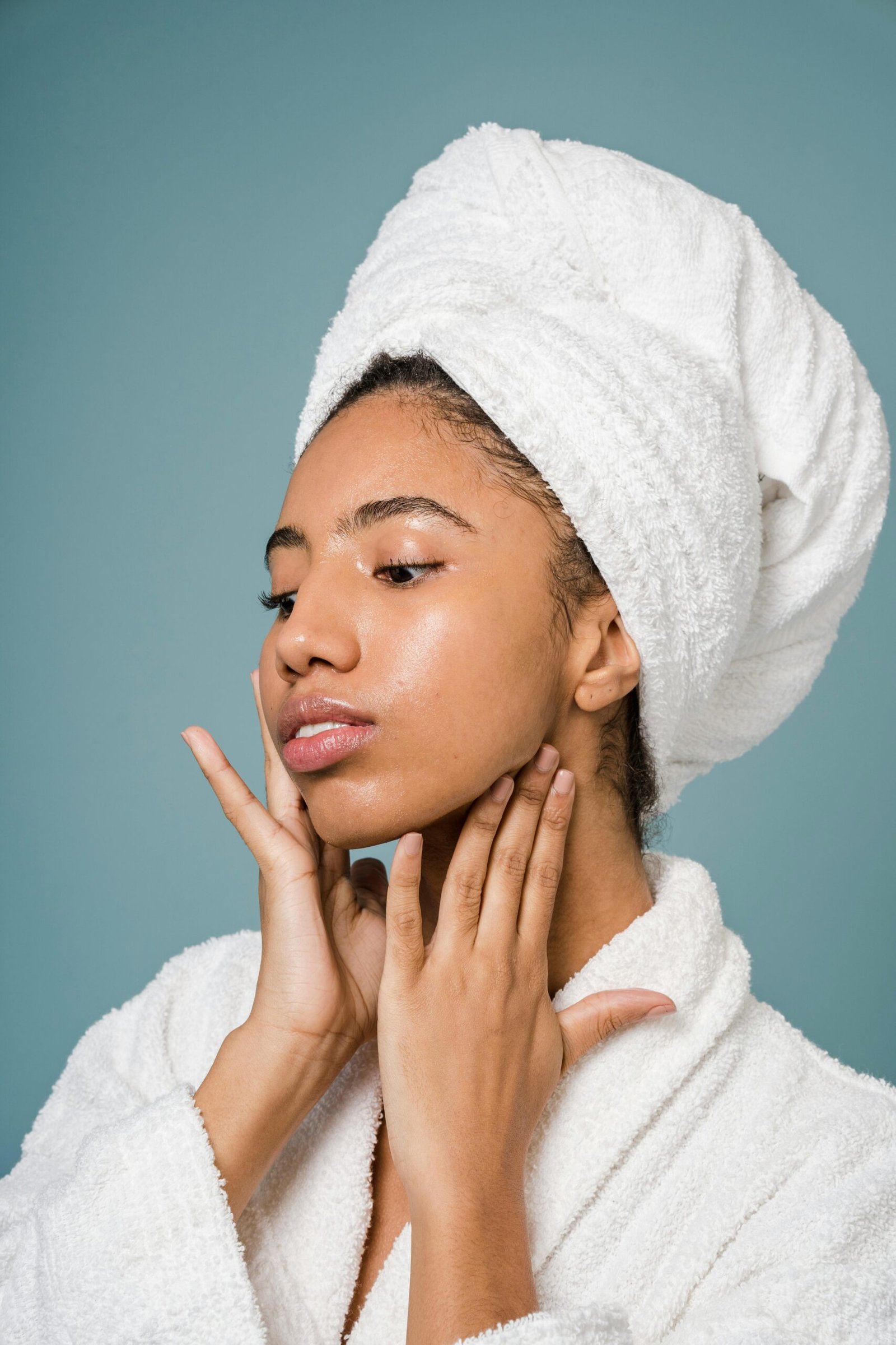A person in a white bathrobe and towel turban touches their neck, set against a soft blue background.
